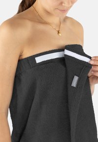 Dunkelgraues Frotteehandtuch mit einer plüschigen Textur. Verfügt über einen weißen Akzentstreifen und ein Etikett an der Seite. Wird gehalten, teilweise geöffnet.