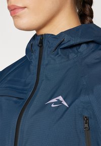 Marineblauwe waterdichte jas met een hoge kraag, zwarte rits en een roze logo. Kenmerken zijn een gestructureerd oppervlak en een verstellbare capuchon.