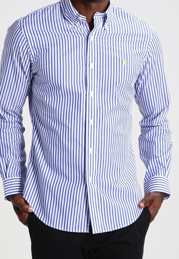 Man wearing a long-sleeve, buttoned blue and white vertical striped shirt with a small yellow logo, hands in black pants pockets.