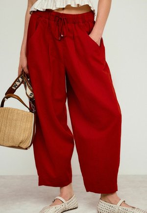 Locker sitzende rote Hose mit elastischem Bund und Kordelzug, getragen mit beigen gewebten Schuhen und einer gewebten Strohtasche mit braunen Ledergriffen.