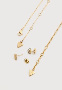 Σετ χρυσού κοσμήματος που περιλαμβάνει κολιέ με αλυσίδα και τριγωνικό μενταγιόν, σκουλαρίκια καρφωτά με διακοσμητική πέτρα, και διάφορα πίσω μέρη σκουλαρικιών.