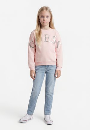 Jauna mergina su ilgais blondinų plaukais, apsirengusi rožiniu megztiniu su sidabrinėmis raidėmis, šviesiai mėlynomis džinsais ir baltais sportbačiais, stovinti baltame fone.