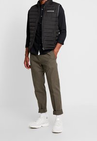 Černá péřová vesta s horizontálním prošíváním, zipem vpředu, bílým logem na hrudi. V kombinaci s olivově zelenými kalhotami a bílými teniskami.