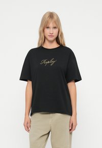 Blond naine, seljas must oversize T-särk, millel on kuldse kiri "Replay" ja beežid püksid, seistes lihtsa valge tausta ees.