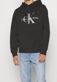 Sudadera negra con bolsillo frontal, que presenta un gráfico de "CK" y "Calvin Klein Jeans" en gris claro. Material de algodón, ajuste relajado, capucha con cordón.