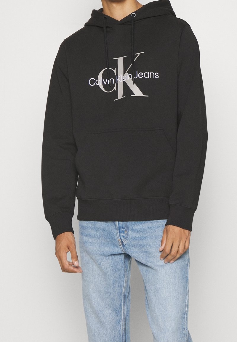 Sudadera negra con bolsillo frontal, que presenta un gráfico de "CK" y "Calvin Klein Jeans" en gris claro. Material de algodón, ajuste relajado, capucha con cordón.
