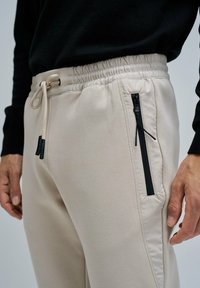 Hombre lleva joggers beige con cintura elástica, cordón y bolsillo negro con cremallera vertical en el muslo derecho, combinados con una camiseta negra.