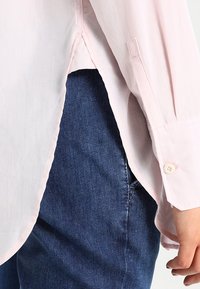 Camicia a maniche lunghe di un delicato rosa chiaro con una texture morbida, dotata di polsini con bottoni e spacchi laterali, abbinata a jeans in denim blu scuro.