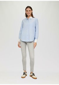 Hellblaues Button-Up-Hemd mit langen Ärmeln und einer Brusttasche, kombiniert mit hellgrauen Skinny-Jeans und zweifarbigen Sneakers mit braunen Sohlen.