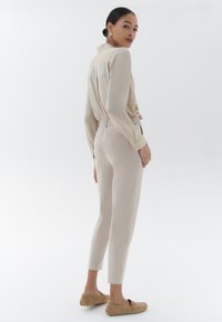 Blouse beige à manches longues avec un col boutonné, associée à un pantalon ajusté beige clair. Le modèle porte des chaussures à enfiler en tan doux.