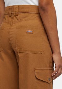 Brown utility pants made from durable fabric, featuring a back pocket with a patch logo, reinforced stitching, and a side cargo pocket.