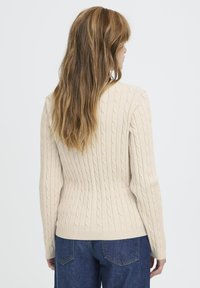 Beige kabelstrikket sweater med lange ærmer og en tætsiddende pasform, der har teksturerede ribmønstre. Båret med mørkeblå jeans.