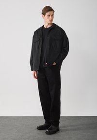 Camisa chaqueta negra de botones con dos bolsillos en el pecho, combinada con pantalones negros y zapatos negros tipo slip-on, con una textura de tela suave.