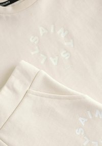 Beige sweatshirt og matchende bukser med et cirkulært hvidt logo med teksten "SAINTS" og "LONDON." Fremstillet af struktureret bomuldsstof.