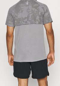 Camiseta deportiva gris con un patrón texturizado, de manga corta y panel trasero sólido en contraste, combinada con pantalones cortos negros.