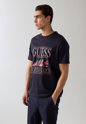 Młody mężczyzna z krótkimi ciemnymi włosami i tatuażem na szyi, ubrany w granatowy t-shirt z napisem "GUESS North Sails" i grafiką żaglówek, stoi z jedną ręką w kieszeni.