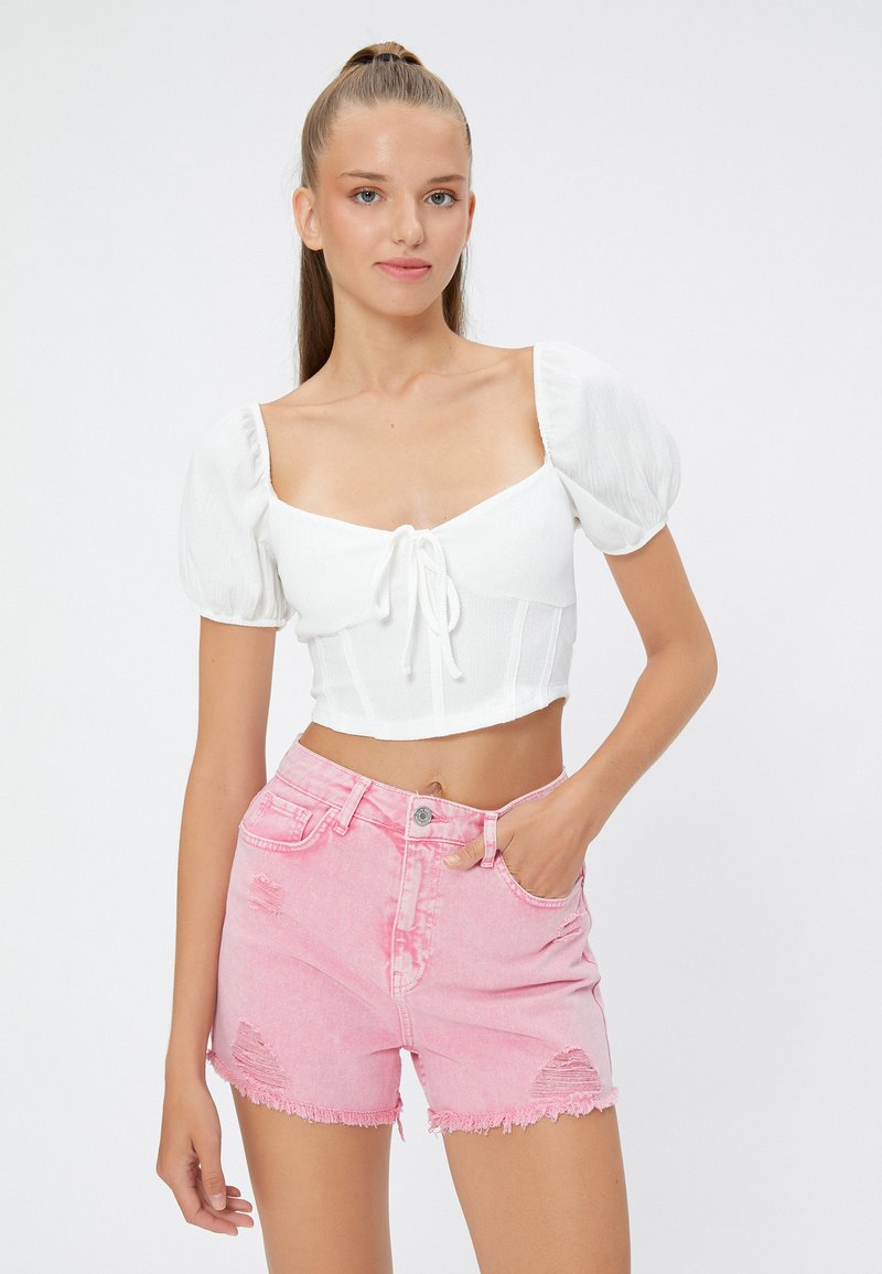 Top corto blanco con mangas abullonadas y detalle de lazo, combinado con pantalones cortos de mezclilla rosados claros con desgaste y dobladillos deshilachados.