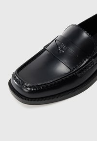 Musta nahkakengät, joissa on kohokuvioitu logo, teksturoidut ompeluseokset, pyöreä kärki ja litteä kumipohja. Sileä pinta ja klassinen muotoilu.