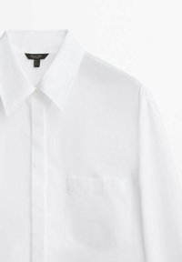 Camicia bianca in cotone con colletto classico, taschino singolo sul petto e chiusura con bottoni sul davanti. Texture liscia e vestibilità sartoriale.