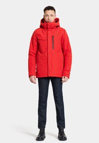 Röd isolerad jacka med hög krage, flera fickor och detaljer i svart dragkedja. I kombination med mörka denimjeans och svarta stövlar.