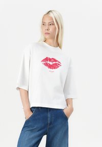 White short-sleeve T-shirt made of soft fabric featuring a red lips graphic and "JOOP!" text at the center. Paired with blue jeans.