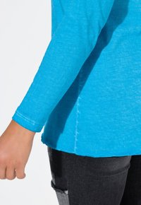 Türkisfarbene Langarmshirt mit abgerundetem Saum, das über eine weiche Textur und sichtbare Nähte verfügt. Kombiniert mit dunkelblauen, ausgewaschenen Jeans.