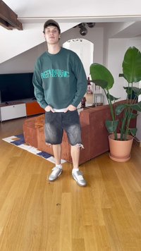 Mladý muž v zelené mikině a černých roztrhaných šortkách stojí v obývacím pokoji s oranžovou pohovkou a velkým květináčem.