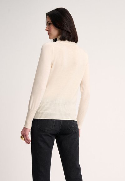 Pulover tricotat de culoare crem, cu o margine și mansete cu model în relief. Designul include un guler înalt și o textură netedă, purtat cu blugi negri.