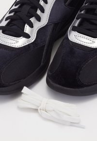 Čierne športové topánky z semišového a sieťovinového materiálu, so striebornými akcentmi na jazyku a bielymi bavlnenými šnúrkami položenými vedľa nich.