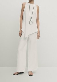 Top bianco senza maniche con orlo asimmetrico abbinato a pantaloni bianchi larghi. L'outfit presenta una lunga collana nera con un pendente circolare.