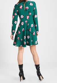 Zelené květinové šaty s dlouhými rukávy a rozšířeným střihem. Obsahují růžové a bílé květinové vzory a detail s klíčovou dírou na zádech.