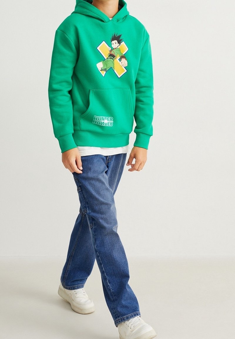 Enfant portant un sweat à capuche vert Hunter x Hunter, un jean bleu et des baskets blanches, marchant sur un fond clair uni.