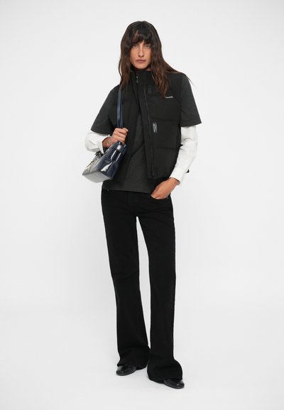 Gilet imbottito nero sopra una maglietta grigia, abbinato a pantaloni neri. Una borsa blu navy e scarpe nere completano l'outfit. Design semplice, vestibilità moderna.
