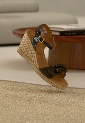 Sandale cu platformă cu talpă înclinată, împletită din iută, curele din piele lac neagră, închideri cu cataramă și talpă căptușită de culoare bej, pe un covor țesut bej.
