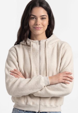 Ung kvinde med langt mørkt hår smiler blidt, iført en lysebeige lynlås-hoodie med hænderne krydset foran på hvid baggrund.