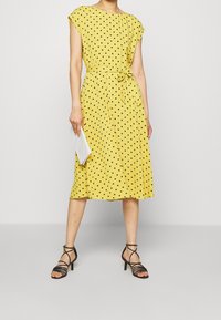 Keltainen pallokuvioinen mekko, jossa on lyhyet hihansuut ja solmittava vyötäröyksityiskohta, yhdistettynä hihattomiin mustiin korkokenkiin ja valkoiseen laukkukoteloon.