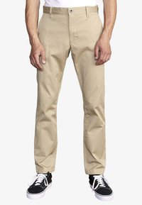 Beige chinos with a straight-leg fit, cotton fabric, belt loops, and a zipper fly. Paired with black sneakers and white laces.