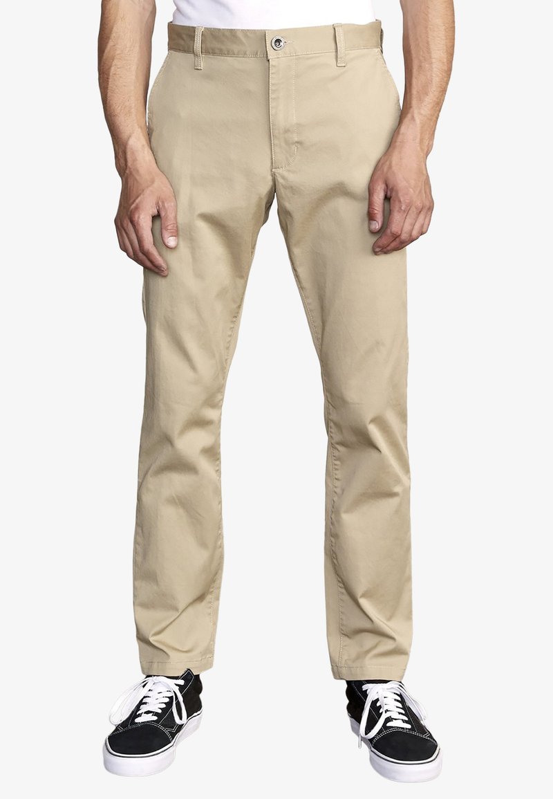 Beige chinos with a straight-leg fit, cotton fabric, belt loops, and a zipper fly. Paired with black sneakers and white laces.