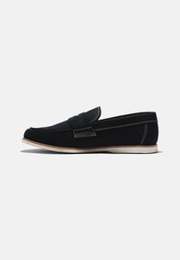 Zapato negro sin cordones con una parte superior texturizada, costuras blancas en contraste y una suela de color claro. Presenta una punta redondeada y un perfil bajo.
