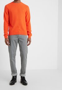 Orangener gerippter Pullover mit langen Ärmeln, kombiniert mit schwarz-weiß gestreiften Hosen. Schwarze Schnürstiefel runden das Outfit ab.