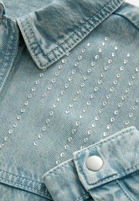 Jasnoniebieski denimiowy materiał z małymi, błyszczącymi ćwiekami ułożonymi w diagonale, z zagiętym mankietem i metalowym zatrzaskiem.