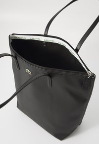 Črna torba iz teksturiranega materiala, z zadrgo, dvema dolgima ročajema in notranjo podlogo s patternom "LACOSTE".