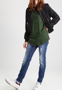 Bomber jacket nera, camicia verde con bottoni, jeans blu strappati e sneakers bianche. I materiali sono tessuto e denim, con una vestibilità casual.