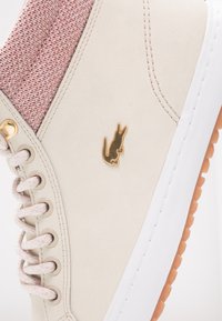 Zapatilla blanca con un cuello textil rosa texturizado, logo dorado y suela de goma marrón claro. Presenta ojales metálicos y cordones con patrón.