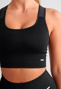 Svart ribbet sportsbh med brede stropper, teksturert stoff og en subtil logo-detalj. Har en tettsittende, cropped design for støtte.