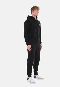 Black hoodie with drawstrings and front pockets, paired with matching black joggers. Notable red logo detail on hoodie. White athletic shoes.