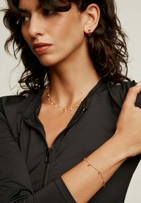 Collar de oro con pequeñas perlas, pulsera de oro a juego y pendientes de aro de oro, todo combinado con una camiseta negra ajustada con cremallera.