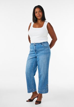 Femme debout portant un haut blanc sans manches, un jean bleu à jambes larges avec des détails de boutons près de l'ourlet, et des talons ouverts noirs.