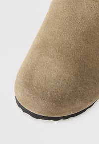 Brown suede ankle boot with a rounded toe and minimalist design. Features a black rubber sole for traction. Soft texture and matte finish.