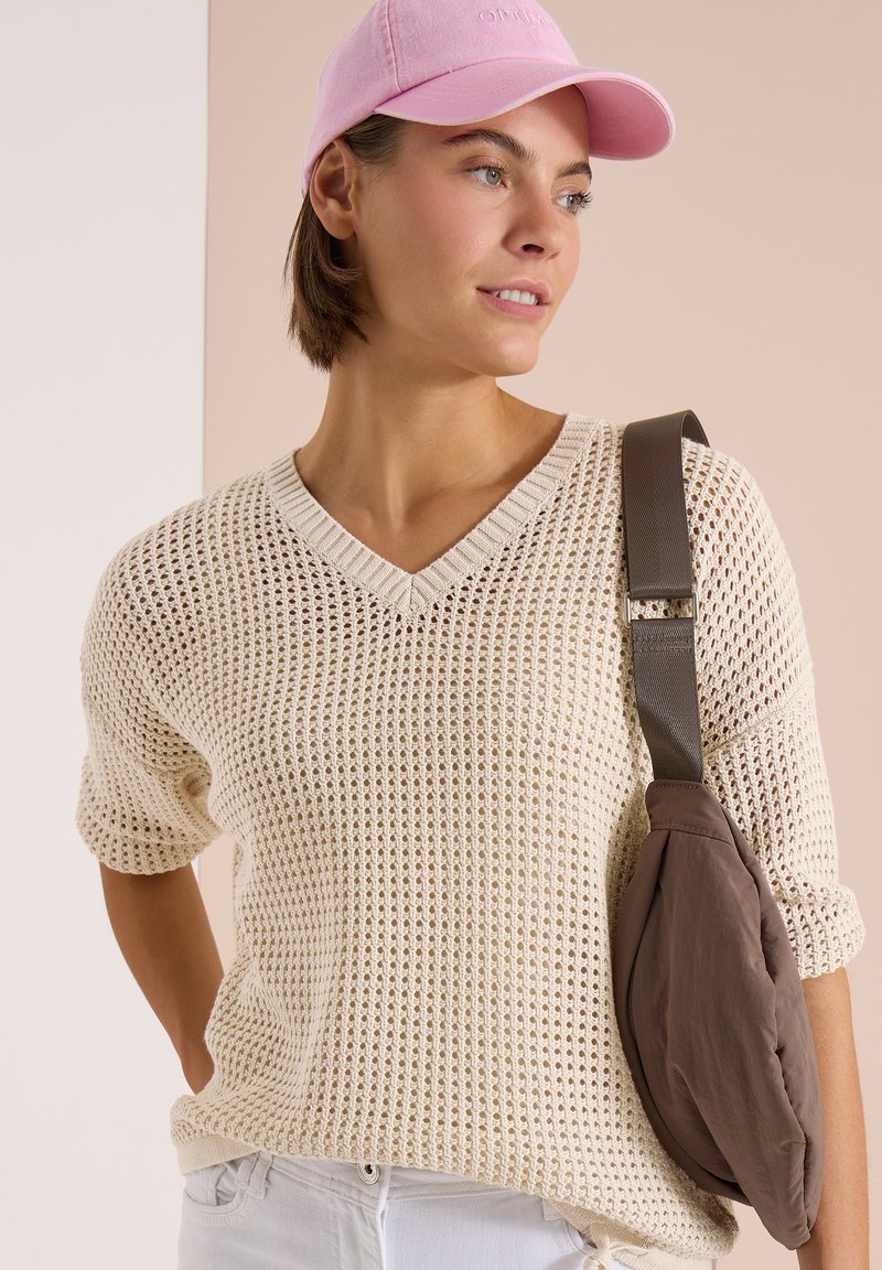 Femme portant une casquette rose, un pull en maille crème à col en V, un pantalon blanc et une bandoulière marron de sac sur l’épaule, regardant sur le côté.